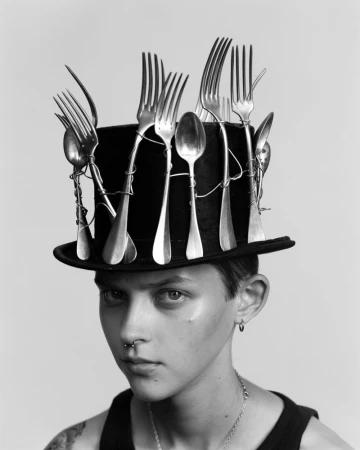 Black and white portrait of a person wearing a black top hat adorned with vintage forks and spoons wired around the crown.