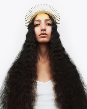 Model with long voluminous crimped dark hair wearing a golden spiked halo crown against a white backdrop.