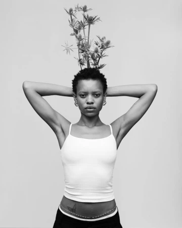 Black and white portrait of a person with thistle flowers atop their short hair, hands behind head in a white tank top.