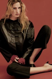 Model Felice Nova Noordhoff seated in a black leather jacket and cropped trousers against a warm terracotta seamless background.