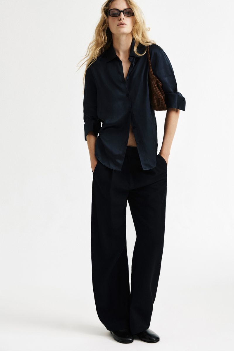 Model wearing dark sunglasses, a navy collared shirt, and black wide-leg trousers against a seamless white studio backdrop.
