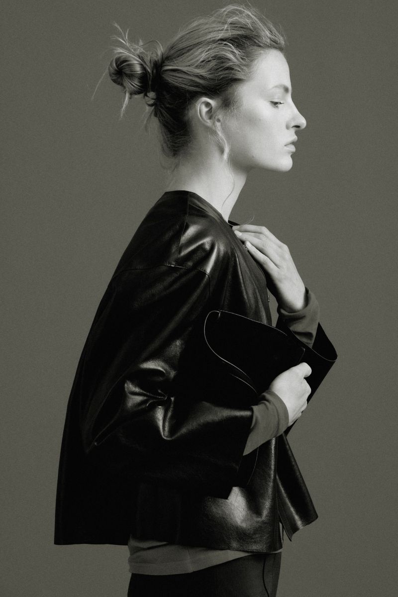 Model Felice Nova Noordhoff in profile wearing a black leather jacket and holding a matching clutch bag against a grey backdrop.
