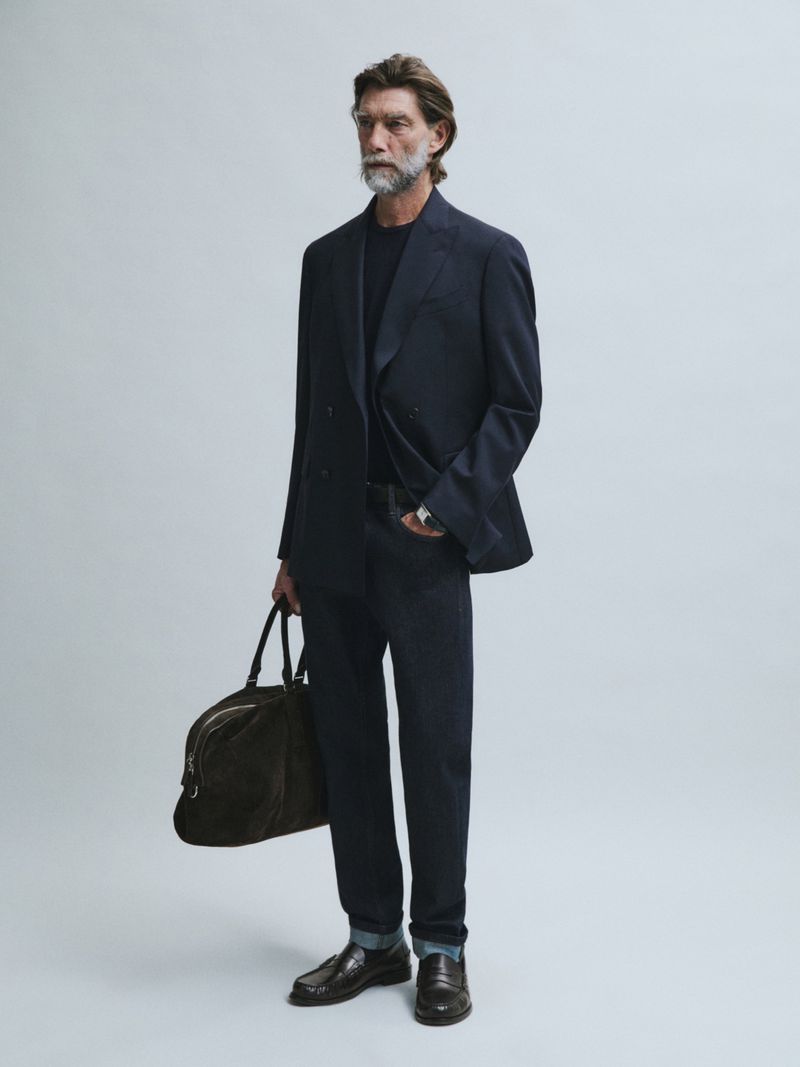Model Patrick Petitjean wearing a navy double-breasted blazer and dark trousers holding a travel bag against a grey background.