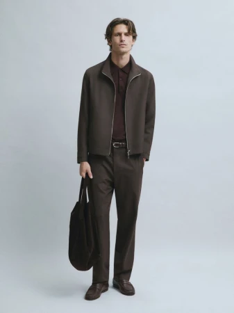 Model wearing a dark brown zip-up jacket, matching trousers, brown loafers, and holding a tote bag against a grey seamless backdrop.