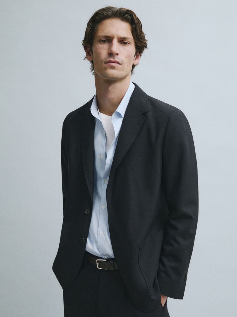 A man with dark brown hair wears a black blazer over a light blue button-down shirt and white undershirt against a light grey background.
