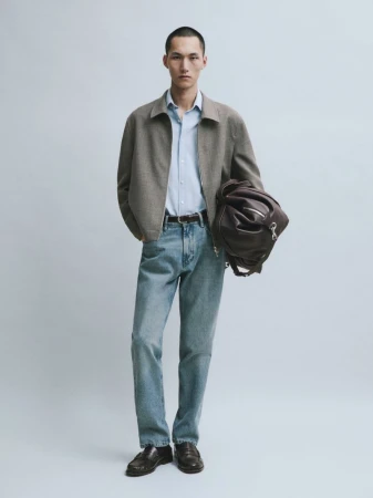 Model Siquan Wang wearing a taupe jacket, light blue shirt, denim jeans, and holding a dark brown leather duffle bag.