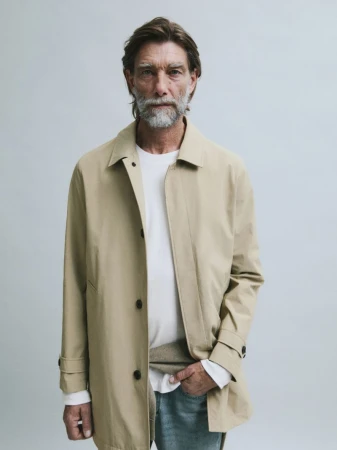 Model Patrick Petitjean posing in a beige overcoat, white shirt, and denim jeans against a pale grey seamless studio backdrop.