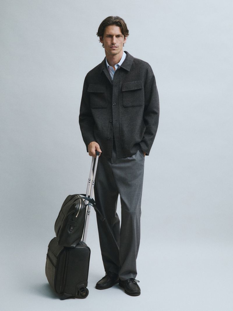 Model Justin Eric Martin in a charcoal jacket and grey trousers holding black luggage against a studio backdrop.
