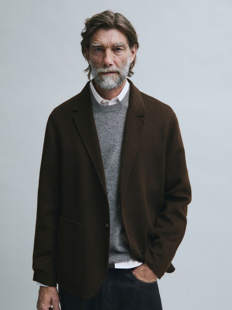 Model Patrick Petitjean wearing a dark brown blazer over a grey sweater against a light grey studio background.