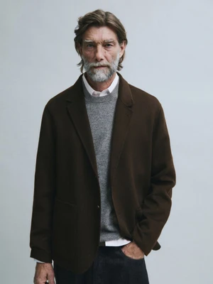 Model Patrick Petitjean wearing a dark brown blazer over a grey sweater against a light grey studio background.