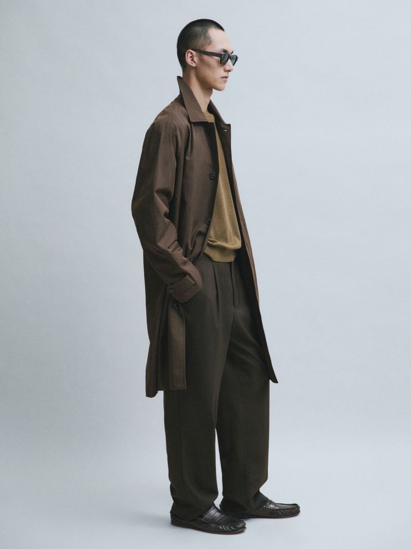 Model Siquan Wang standing in profile wearing a brown trench coat, olive trousers, and sunglasses against a grey studio backdrop.
