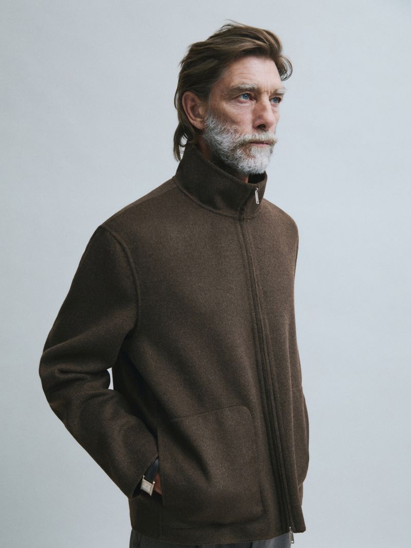 Model Patrick Petitjean wears a dark brown zip-up jacket against a light grey studio backdrop in a profile portrait.