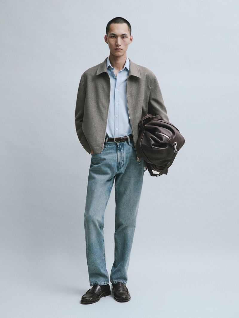Model Siquan Wang wearing a taupe zip-up jacket, light blue shirt, straight-leg blue jeans, and holding a brown leather duffle bag.