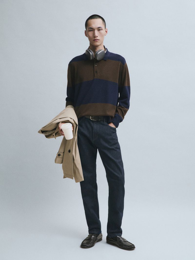 Model Siquan Wang standing in a striped knit polo, dark trousers, loafers, and headphones holding a coat for Massimo Dutti.