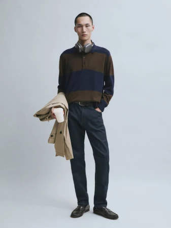 Model Siquan Wang standing in a striped knit polo, dark trousers, loafers, and headphones holding a coat for Massimo Dutti.