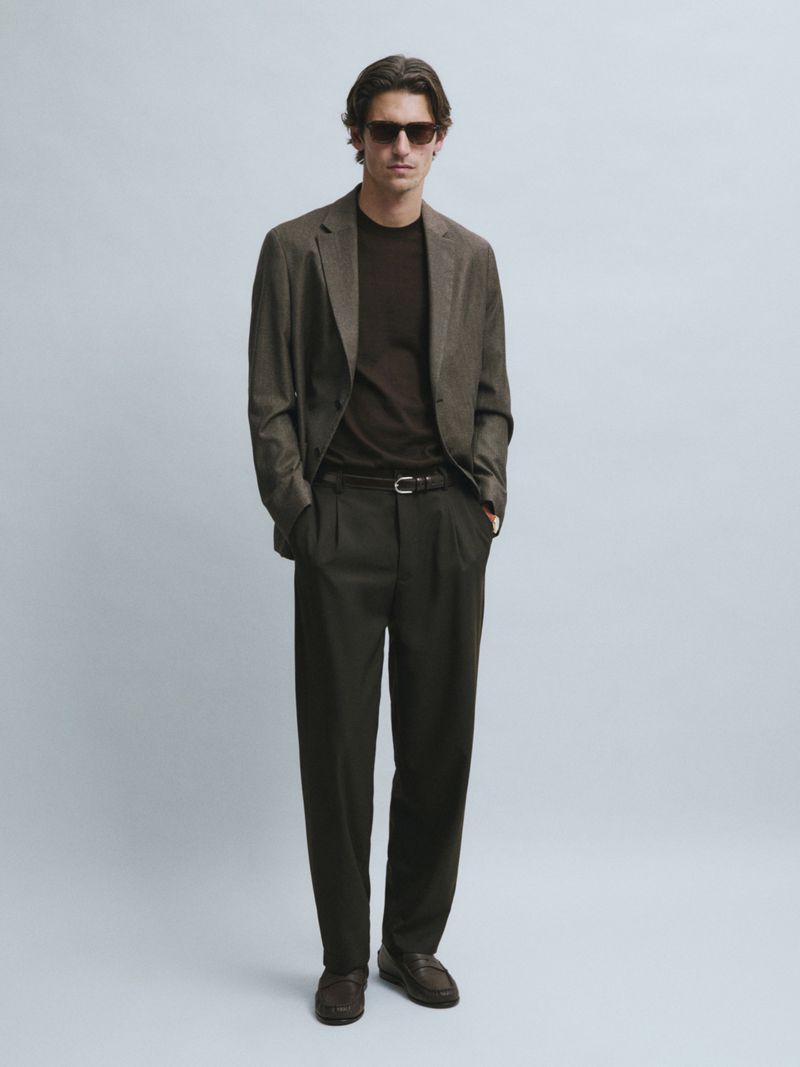 Male model in dark sunglasses, tailored olive-brown blazer, and pleated trousers against a pale grey studio background.