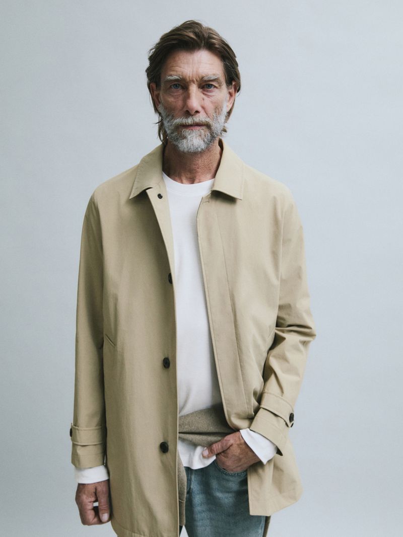 Model Patrick Petitjean wears an unbuttoned beige mac coat over a white top and faded jeans against a light grey studio background.