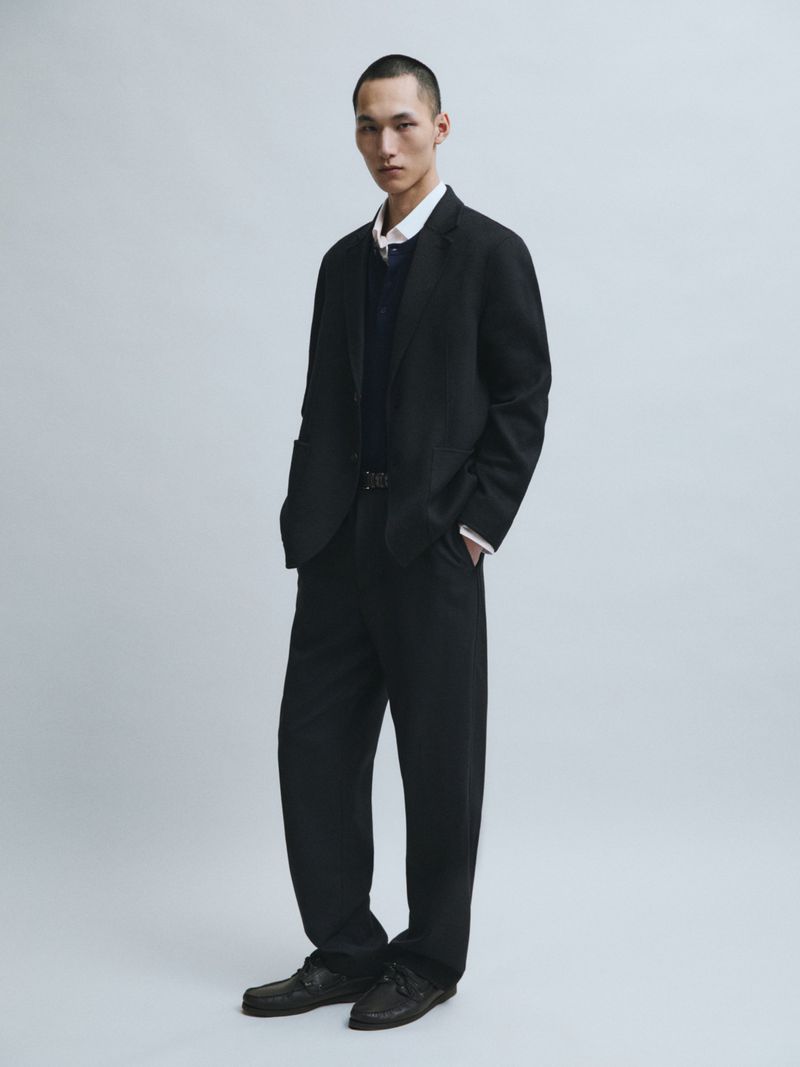 Model Siquan Wang standing in a relaxed black suit, collared shirt, and dark leather shoes against a grey studio backdrop.