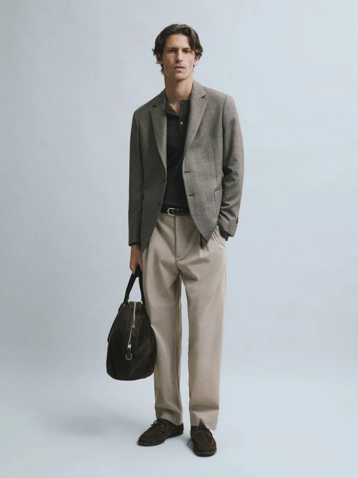 Man standing in a tailored grey blazer and pleated beige trousers holding a dark brown suede bag against a plain studio backdrop.