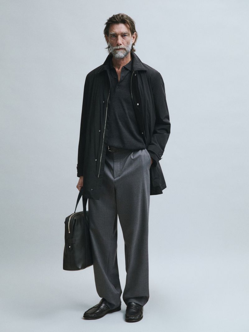 Model Patrick Petitjean wearing a black jacket, grey trousers, and holding a black leather bag against a neutral studio backdrop.