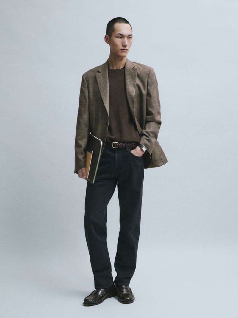 Siquan Wang wears a brown blazer, knit sweater, dark trousers, and leather loafers while holding a document folder against a grey backdrop.