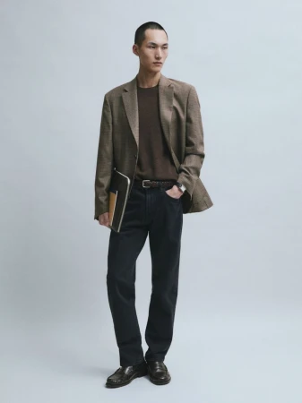 Siquan Wang wears a brown blazer, knit sweater, dark trousers, and leather loafers while holding a document folder against a grey backdrop.
