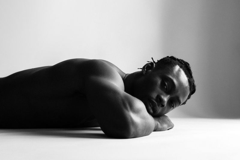 Monochrome portrait of a muscular Black adult male lying down in a studio, showcasing fitness and skin texture against a light background.