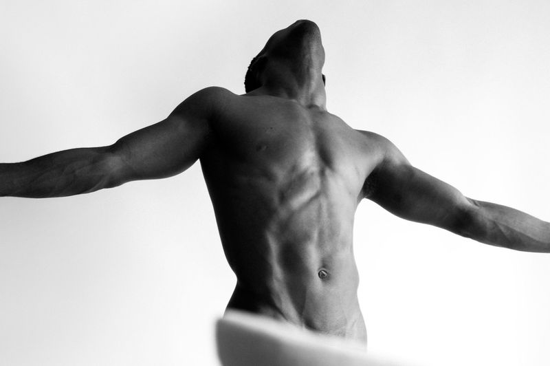 Black and white studio shot of a muscular adult male with his arms outstretched, head tilted back, and torso visible against a white backdrop.