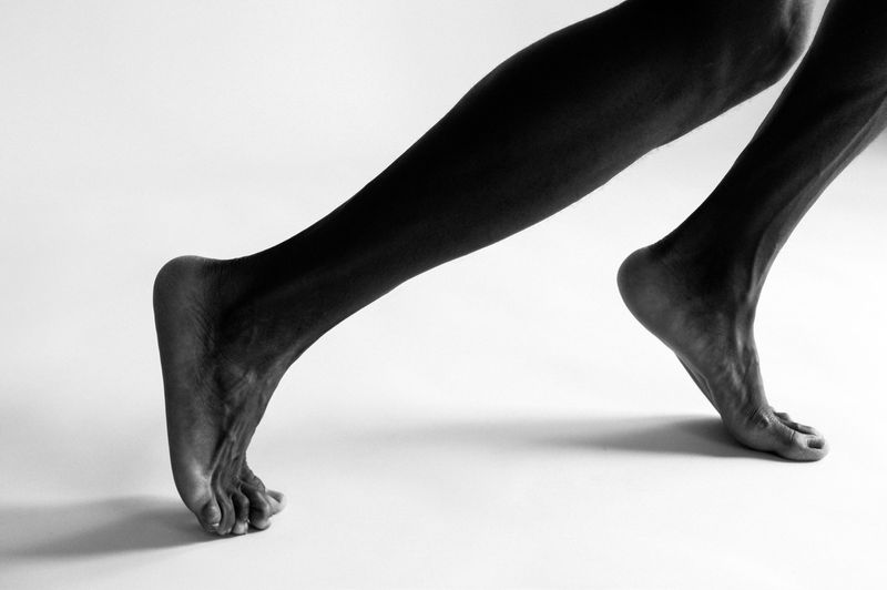 Close-up studio shot of the lower body of a dark-skinned person walking. Focus on feet and legs against a clean white background.