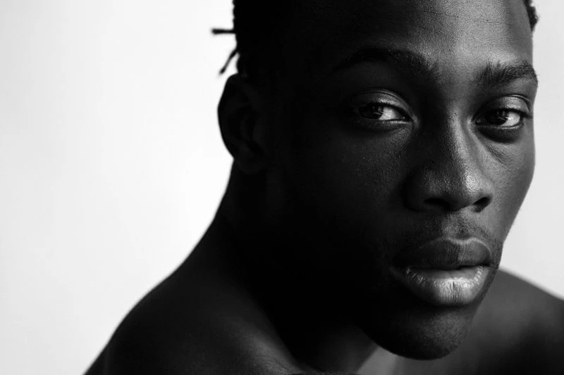 Striking black and white portrait of a dark-skinned male model with short dreadlocks against a stark white backdrop. Focus on facial details.