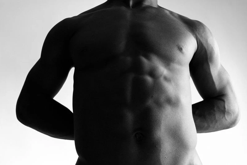 Black and white studio shot of a muscular adult male torso and arms against a gradient white backdrop with neutral facial expression.
