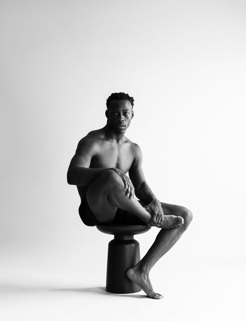 Monochrome image of an athletic, shirtless Black man sitting on a black stool with legs crossed in a white photo studio.