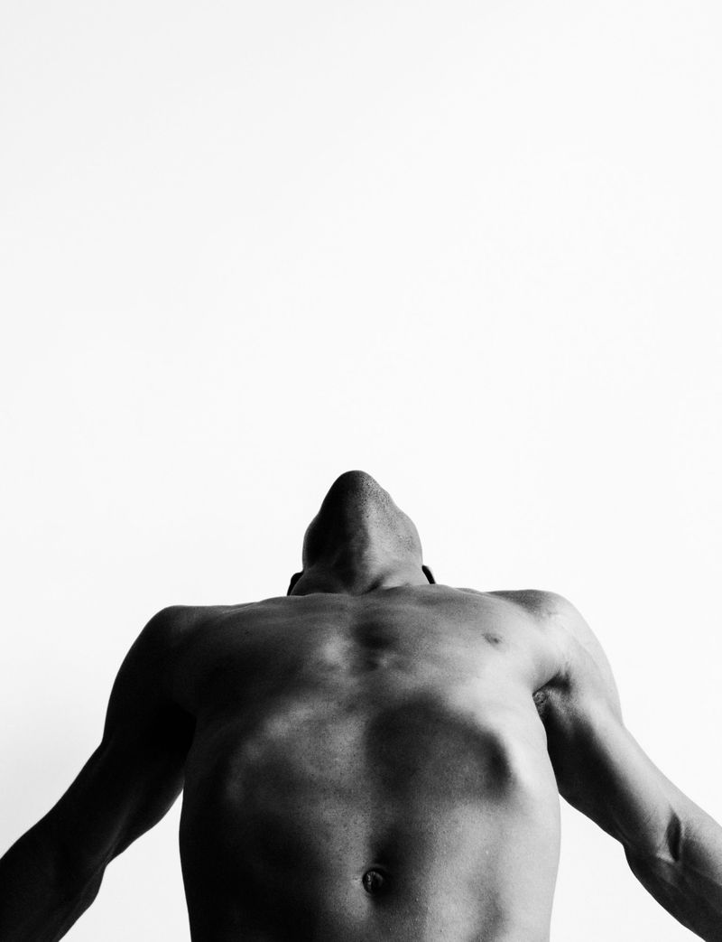 Black and white portrait of an adult male with muscular physique, looking up with arms extended, set against a stark white backdrop.