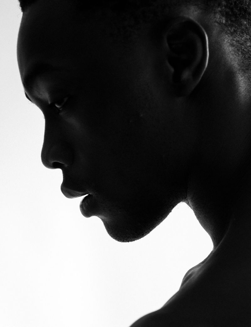 High-contrast black and white side profile portrait of a Black man in deep shadow against a bright white background.
