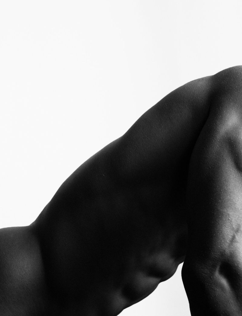 Monochromatic studio shot of the upper torso and leg of a muscular adult male in a bent over pose against a white backdrop.