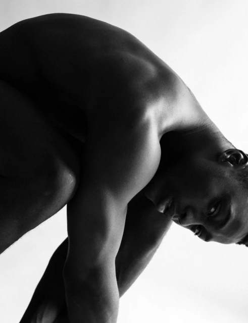 Monochrome portrait of a muscular, dark-skinned adult male in a contorted pose with a white background, showcasing fitness and flexibility.