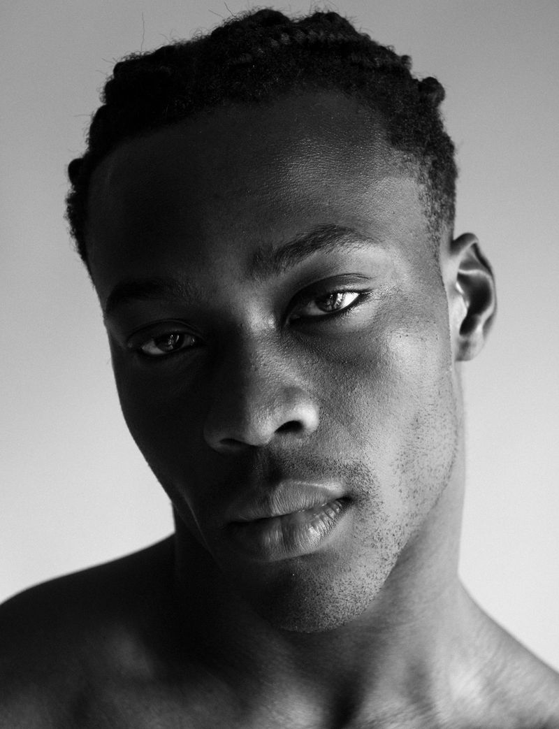 Monochrome portrait of a young Black man with braided hair, captured in a close-up shot with a neutral expression against a light background.