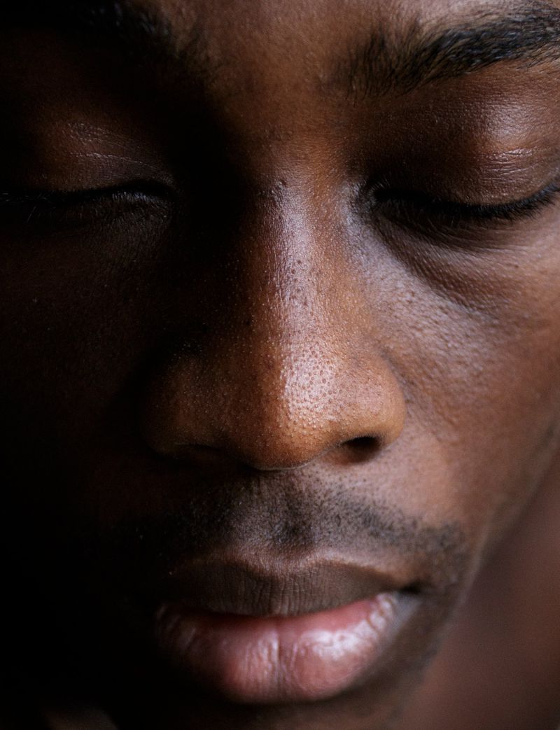 Close-up portrait of an adult African American male with closed eyes, showcasing dark skin tones and textures under soft, directional lighting.