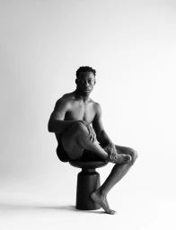 Black and white portrait of a shirtless Black man sitting on a dark sculptural stool against a white studio background.