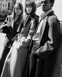 Three models wearing tailored wool coats and outerwear posing outdoors against a sunlit building wall in black and white.