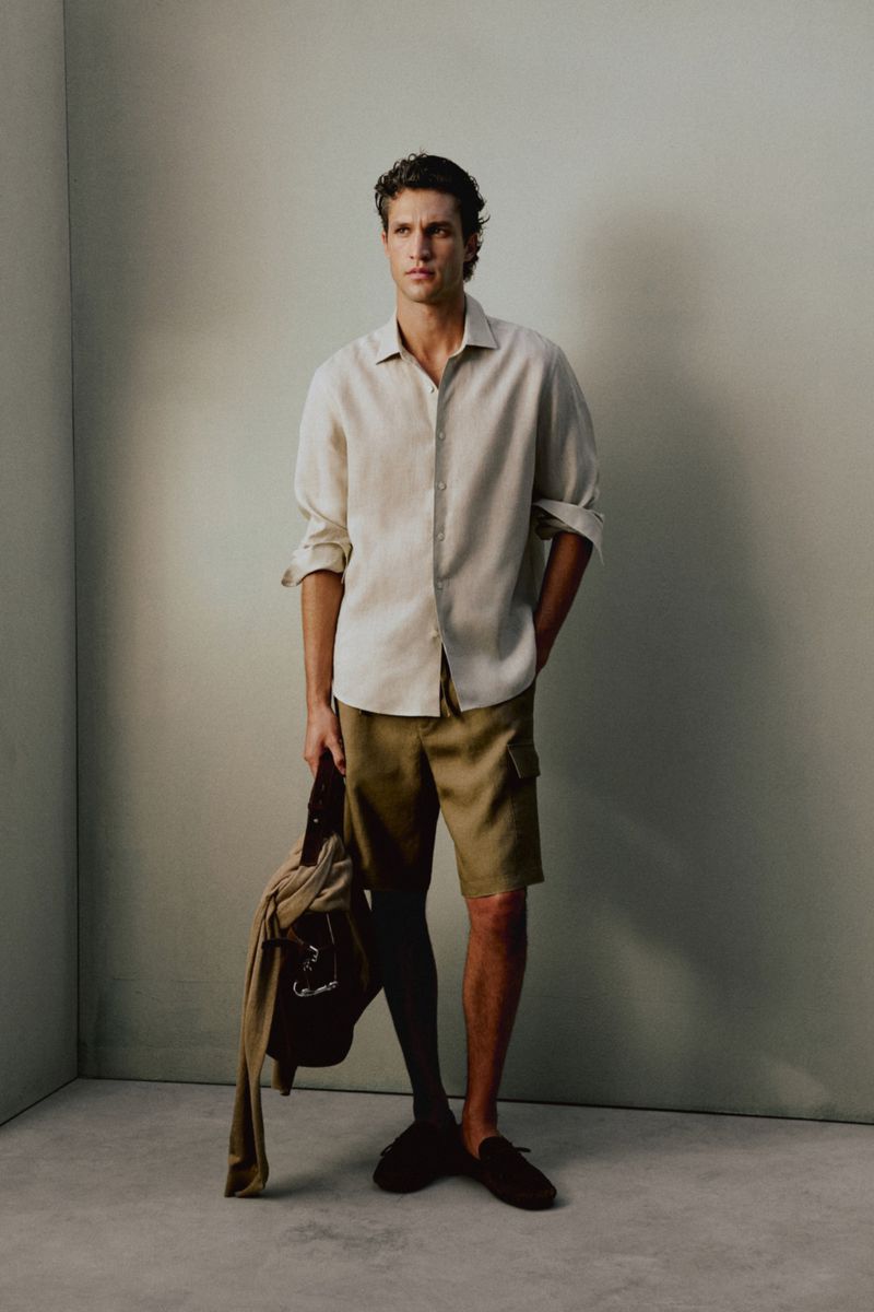 Model Francisco Henriques wearing a cream linen shirt and olive shorts holding a bag in a studio setting for Massimo Dutti.