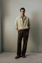 Model Francisco Henriques standing in a cream linen button-up shirt and dark pleated trousers against a neutral grey wall.