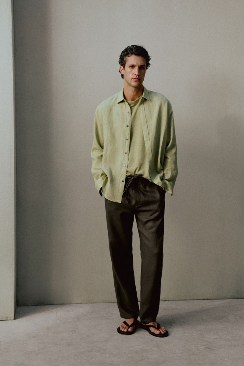 Model Francisco Henriques wearing a light sage shirt and dark relaxed trousers against a neutral grey studio backdrop.