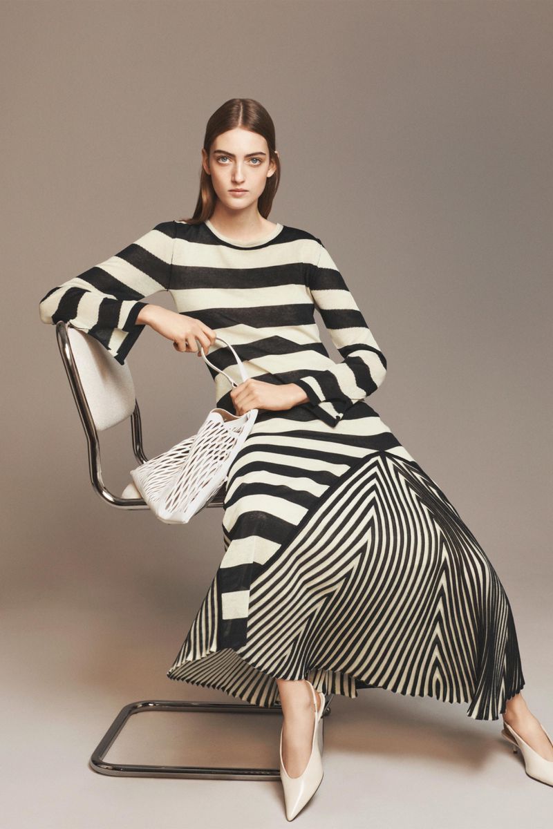 Model wearing a black and white striped dress with a flared skirt seated on a modern chrome chair holding a white mesh handbag.