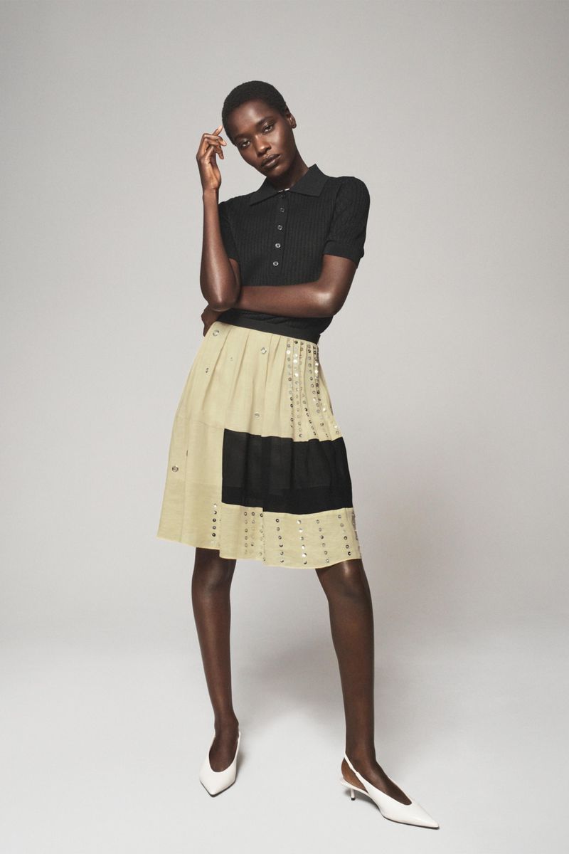 Model Awar Odhiang wearing a black polo knit shirt, embellished beige pleated skirt, and white slingback heels on a studio seamless backdrop.