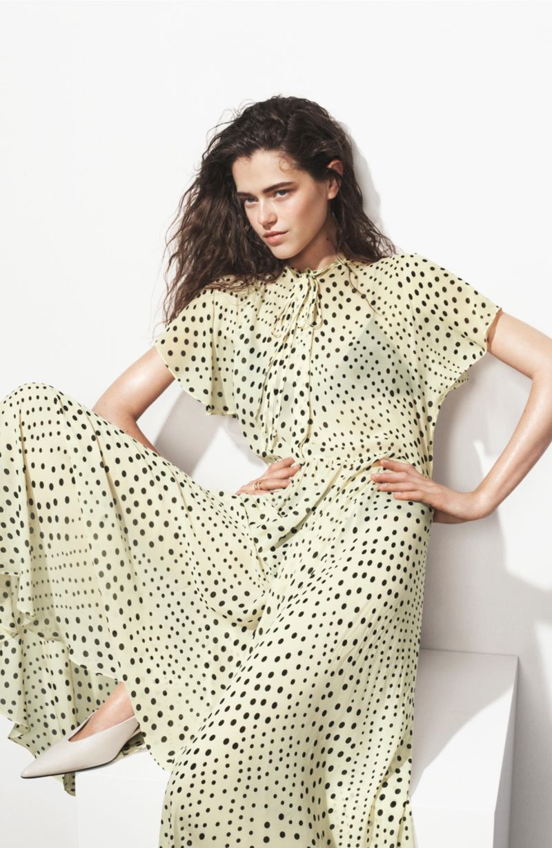 Model wearing a pale yellow polka dot maxi dress seated on a white block against a white studio backdrop.