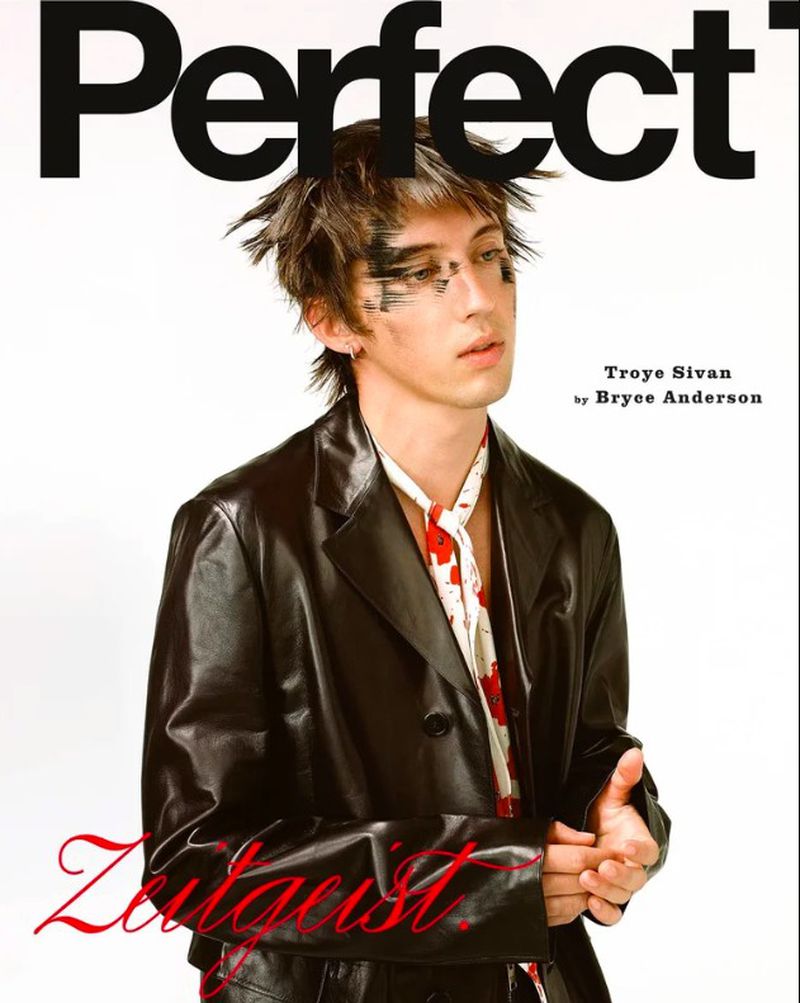 Troye Sivan with messy dark hair and black eyeliner makeup wears a black leather jacket and white patterned scarf, against a plain white background