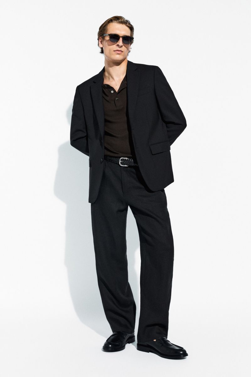 Colin Otto standing against a white backdrop wearing a black blazer, dark polo shirt, black trousers, and sunglasses.