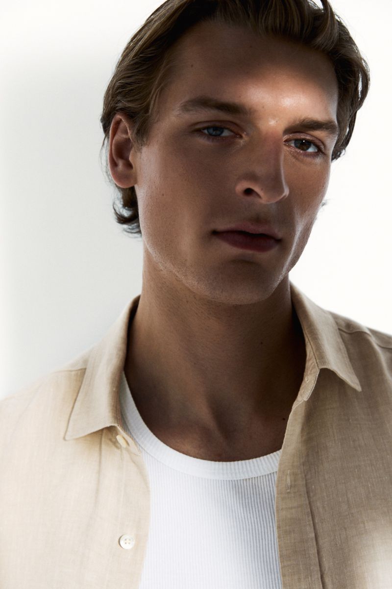 Close-up of a man wearing an open beige linen shirt over a white ribbed tank top, lit with high-contrast shadows against a white backdrop.