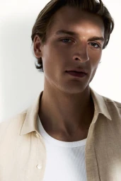 Model Colin Otto wearing an open beige linen shirt over a white ribbed tank top against a white background.
